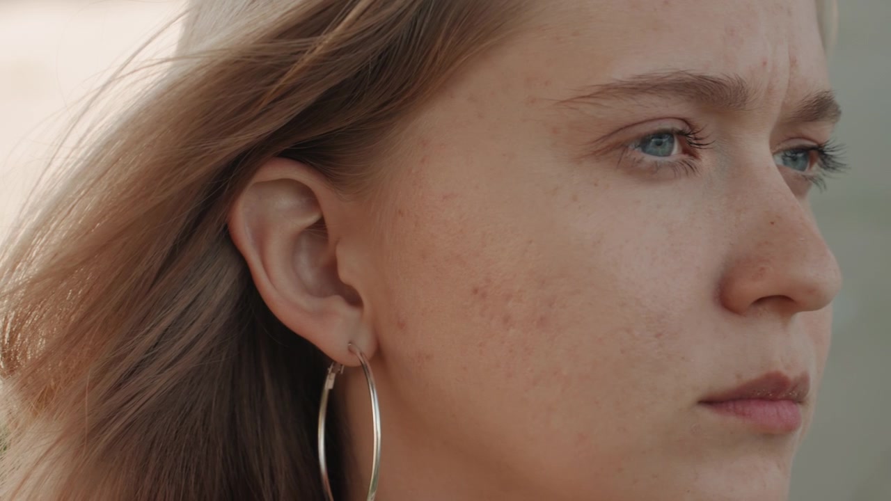 Vista ravvicinata dell'orecchio di una donna e dei capelli lunghi con illuminazione soffusa che evidenzia i dettagli della texture. I capelli si muovono delicatamente nella brezza mentre l'orecchino rimane visibile. Lo sfondo è leggermente sfocato.