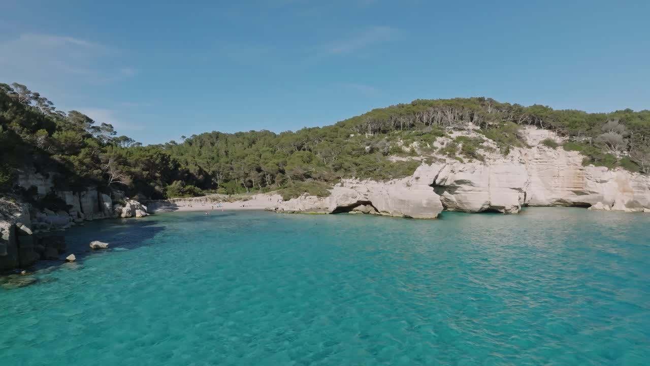 la playa de arena blanca de cala virgen reluce mientras un avión no tripulado vuela sobre el agua en un claro día de verano en menorca, españa.