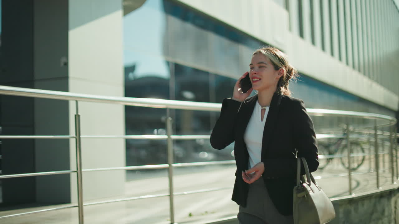Professional business lady walking home smiling during phone call, dressed in formal attire, holding handbag, enjoying conversation under bright sunlight, urban glass building