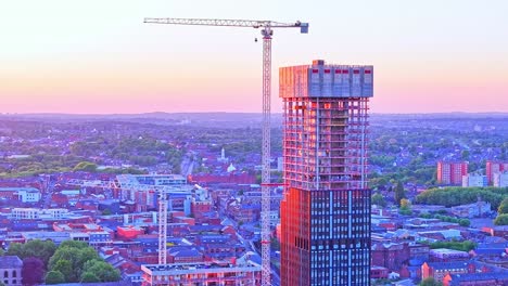 drone footage shows a skyscraper in construction, with cranes in the birmingham city center near pigeon park, as sunset light bathes the urban landscape in warm colors