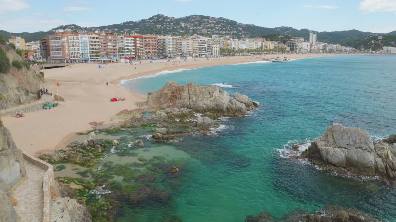 Vista della spiaggia principale di Lloret de Mar con gli edifici sullo sfondo e l'acqua trasparente turchese della spiaggia registrata in 4K 10bit