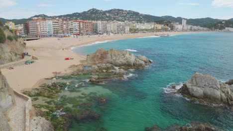 Vista-De-La-Playa-Principal-De-Lloret-De-Mar-Con-Los-Edificios-Al-Fondo-Y-El-Agua-Azul-Turquesa-Transparente-De-La-Playa-Grabada-En-4k-10bit