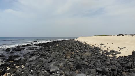 Lufttiefflug-über-Dunkle-Felsen,-Der-Neben-Dem-Sandstrand-Von-Kap-Verde-Einen-Wellenbrecher-Bildet