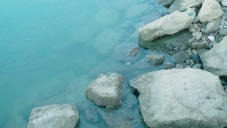 Tilt-Up-of-Plastic-Jug-and-Litter-Pollution-on-Rocks-at-Edge-of-Water