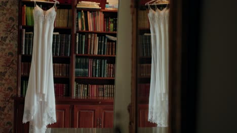 a white wedding dress hanging in front of a bookshelf filled with colorful books in a cozy, dimly lit room