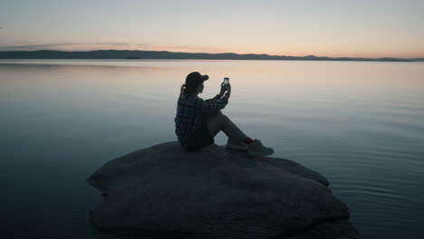 Mujer-Sentada-En-Una-Roca-En-El-Lago-Y-Fotografiando-La-Puesta-De-Sol-Con-El-Teléfono