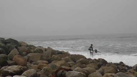 Surfista-Parado-En-La-Costa-Rocosa-Del-Océano-Mientras-Se-Prepara-Para-Remar-Hacia-El-Mar