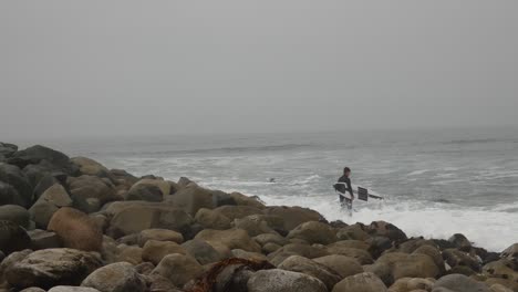 surfer standing on rocky ocean shore as he gets ready to paddle out to sea