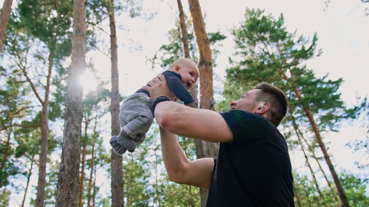Handsome muscular Caucasian man tossing his little baby boy. Spending time with child in nature. Low angle view. Pine trees at backdrop.