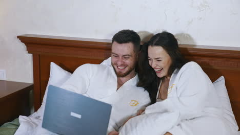 joyful young couple on bed. handsome man and attractive woman in white bathrobes laying on bed and laughing while looking into laptop. happy together.