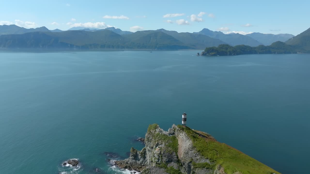 Drone footage of a striped lighthouse on a rocky green headland above calm blue sea, with mountain ranges on the horizon in Vilyuchinskaya Bay, Kamchatka Krai, Russia