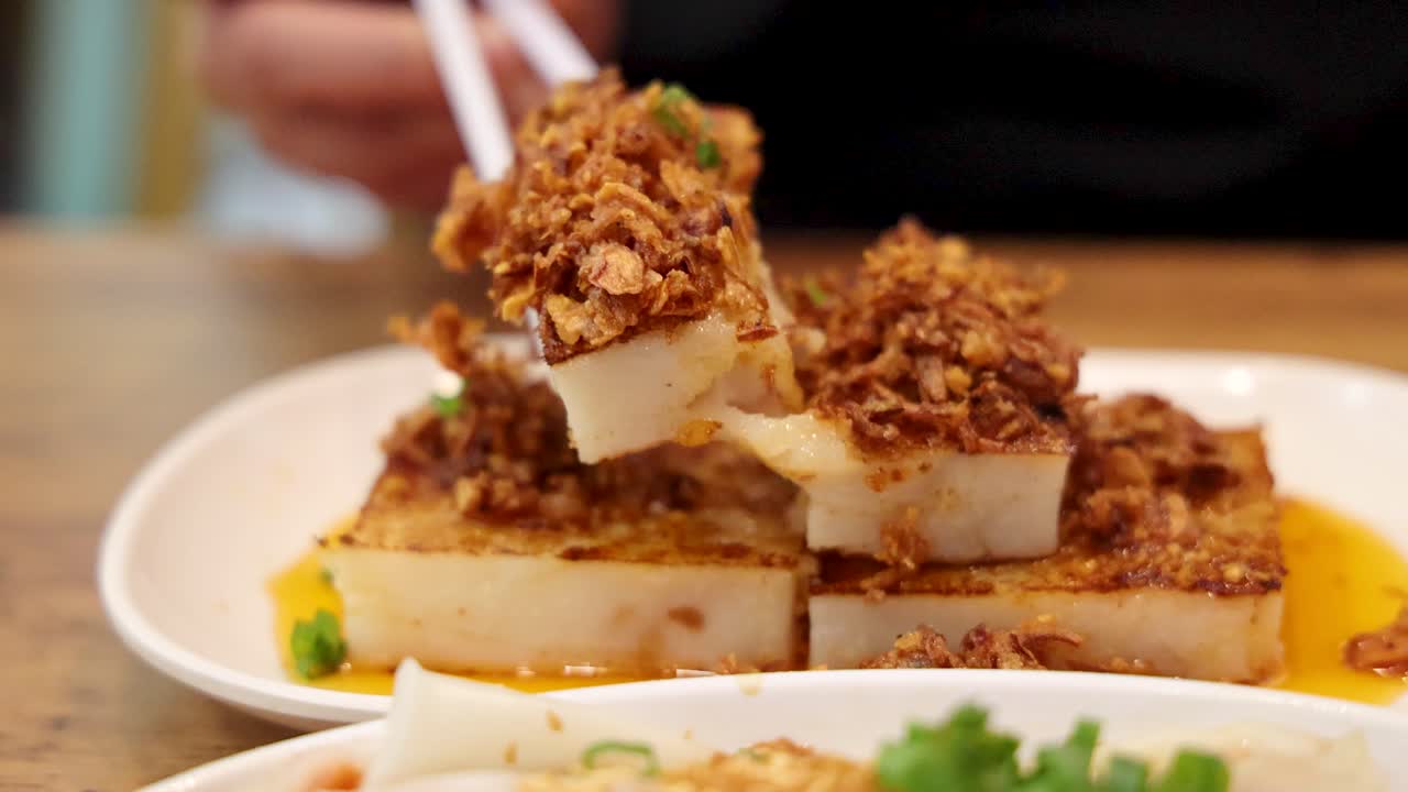 Close-up of chopsticks picking up a piece of fried turnip cake topped with savory garnish in a well-lit Singaporean restaurant, shallow depth of field