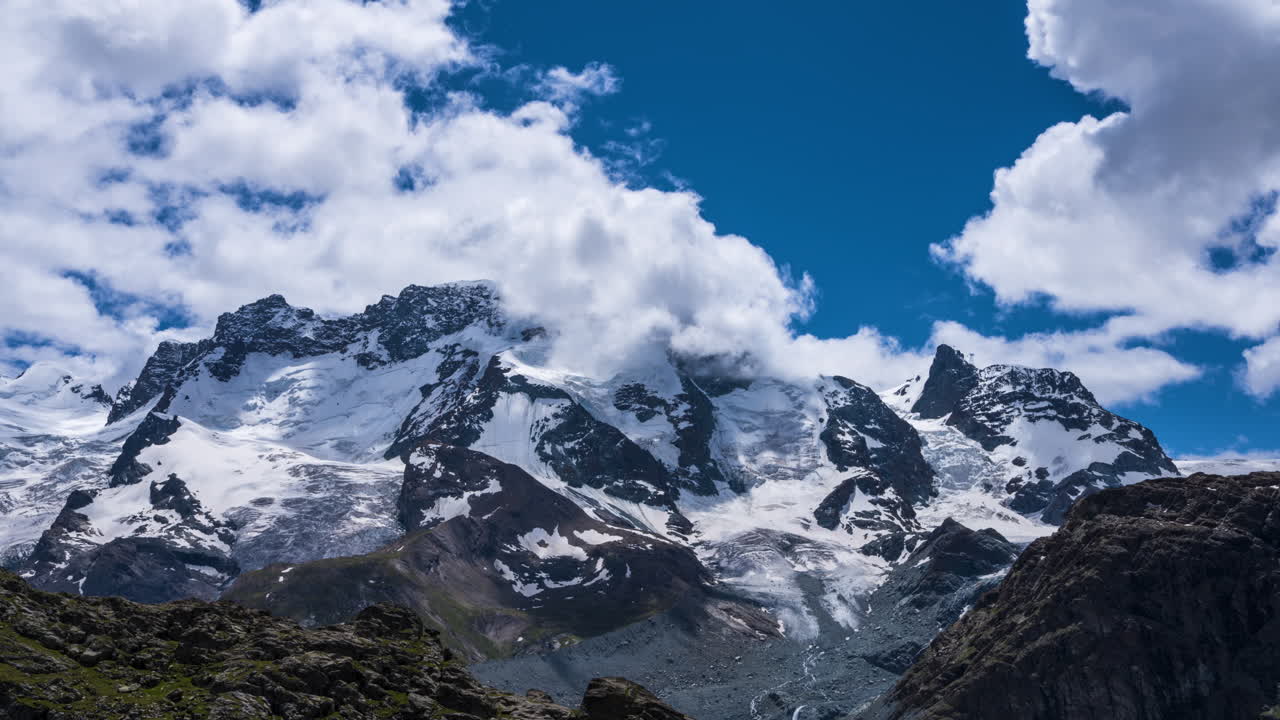 esponjosas nubes blancas rodando contra el cielo azul sobre la cumbre del macizo cubierto de nieve en breithorn cerca de zermatt, suiza - lapso de tiempo