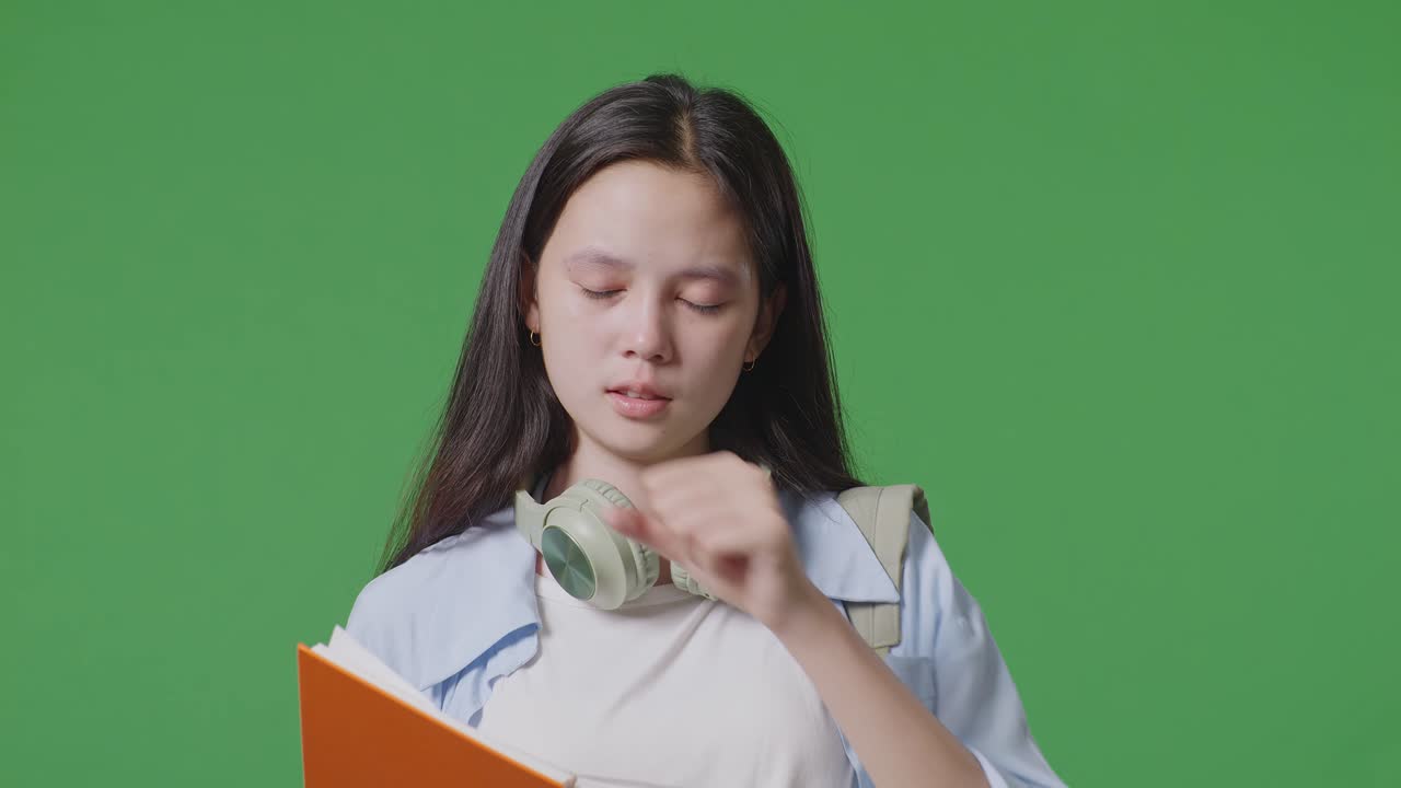 close up de una estudiante adolescente asiática con una mochila leyendo un libro y bostezando mientras está de pie en el estudio de fondo de la pantalla verde