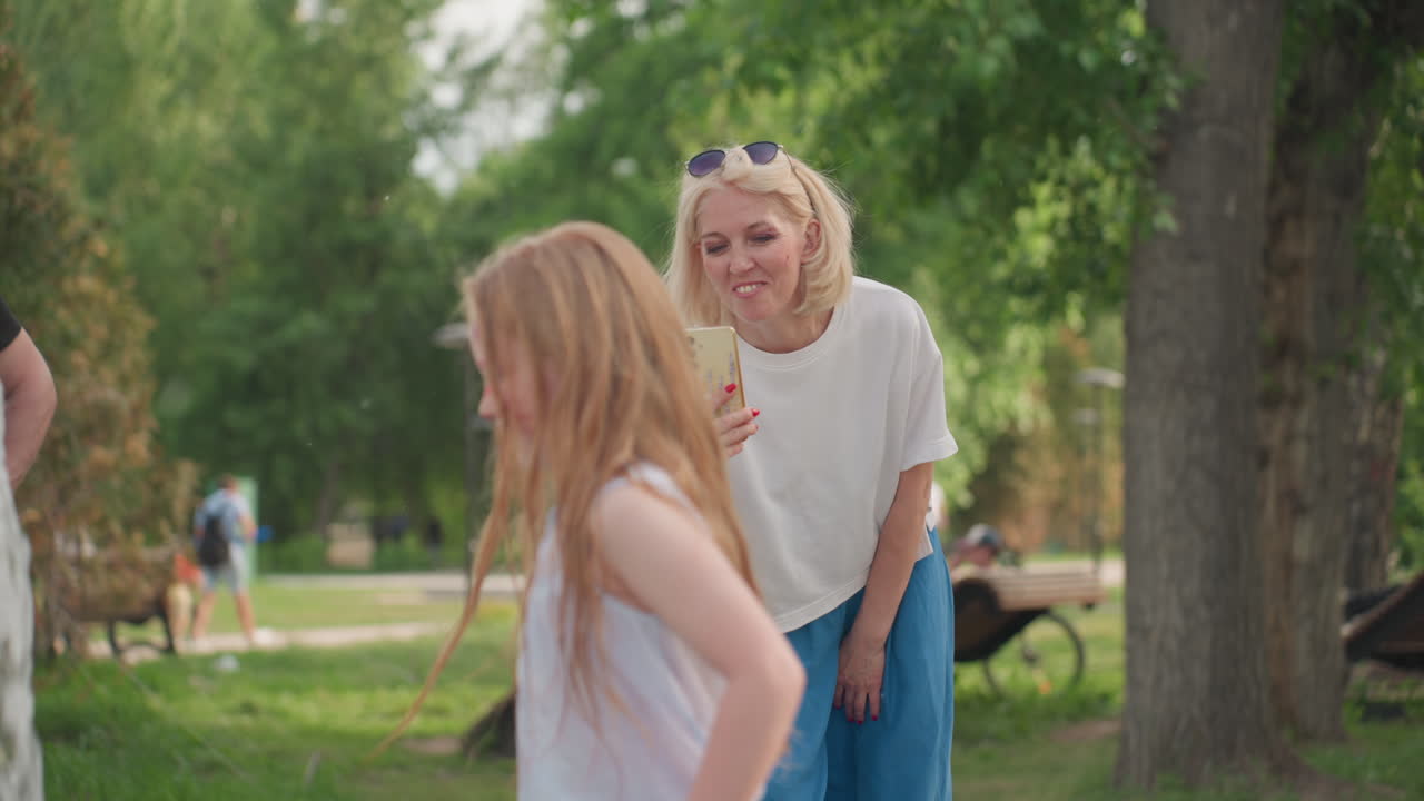 mom in summer outfit holding phone filming daughter playing near fountain, joyful interaction captured in green park, candid family moment full of laughter and movement, warm outdoor bonding memory