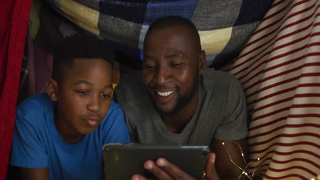 Excited-african-american-father-and-son-using-tablet-lying-in-blanket-camp