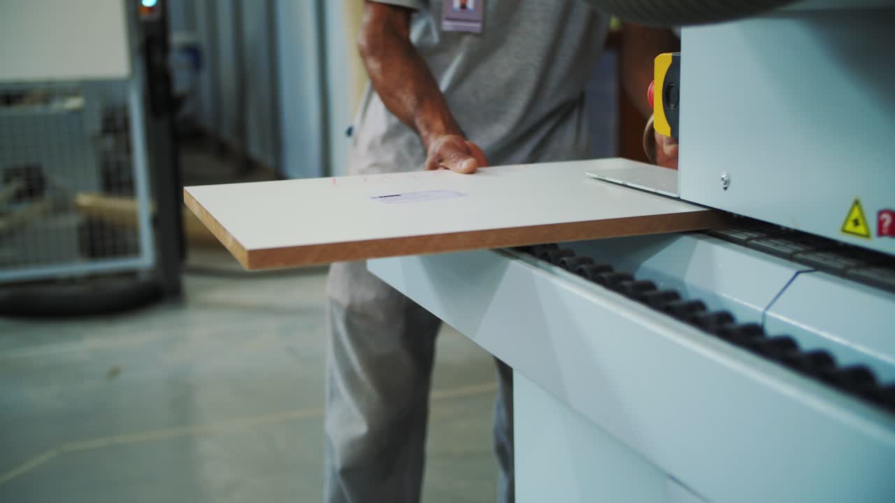 The image shows a person feeding a white wooden board into an industrial machine for processing. The machine has safety markings, and the workshop environment in the background is slightly blurred.