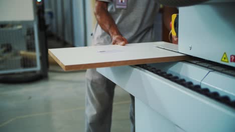 the image shows a person feeding a white wooden board into an industrial machine for processing. the machine has safety markings, and the workshop environment in the background is slightly blurred.