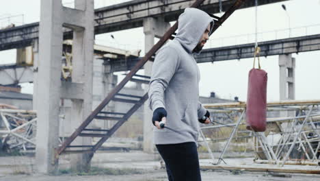 hombre caucásico con barba en una sudadera con capucha gris saltando la cuerda al aire libre en una fábrica abandonada en una mañana nublada