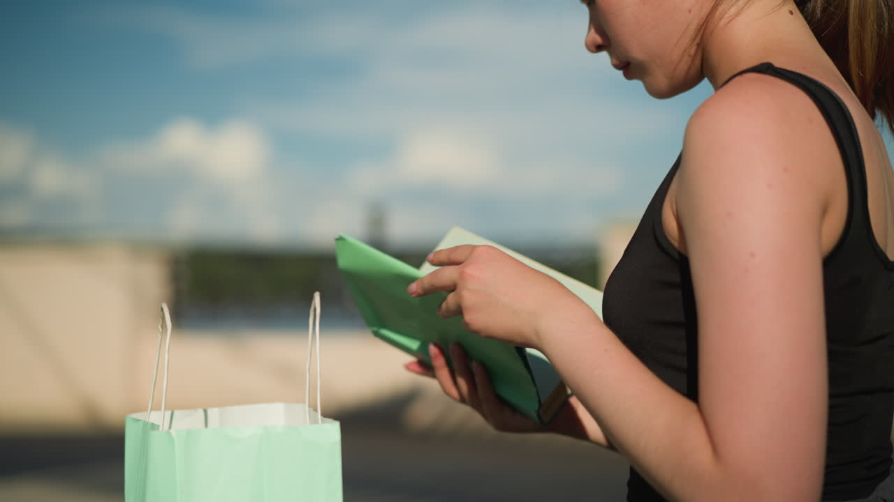 야외 벤치에 앉아있는 여성의 클로즈업, 민트 녹색 가방에서 책을 가져오고, 그녀는 책을 열고 페이지를 뒤집습니다. 흐릿한 배경에는 울타리와 먼 다리가 있습니다.
