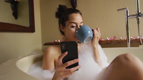 Relaxed-biracial-woman-with-vitiligo-taking-bath-with-foam,-drinking-coffee-and-using-smartphone