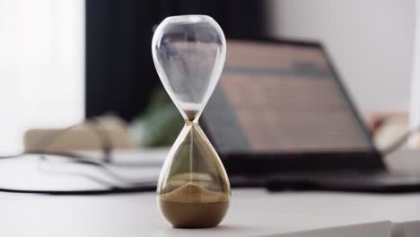 sand clock or hourglass, office and laptop in background, closeup view