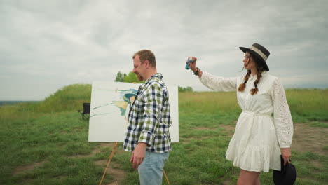 a woman in a stylish hat and white dress sprays mosquito repellent on a man s head in a grass field. the man, in a checkered shirt, stands calmly as she sprays. a painting board and chair are visible