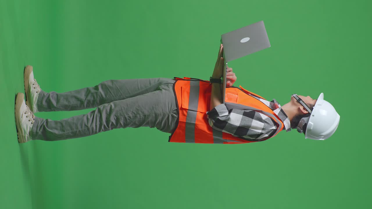 Full Body Side View Of Asian Male Engineer With Safety Helmet Working On A Laptop And Looking Around While Standing In The Green Screen Background Studio