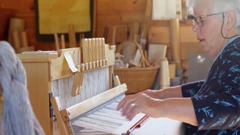 Side-view-of-old-caucasian-senior-woman-weaving-cloth-on-handloom-machine-in-a-workshop-4k