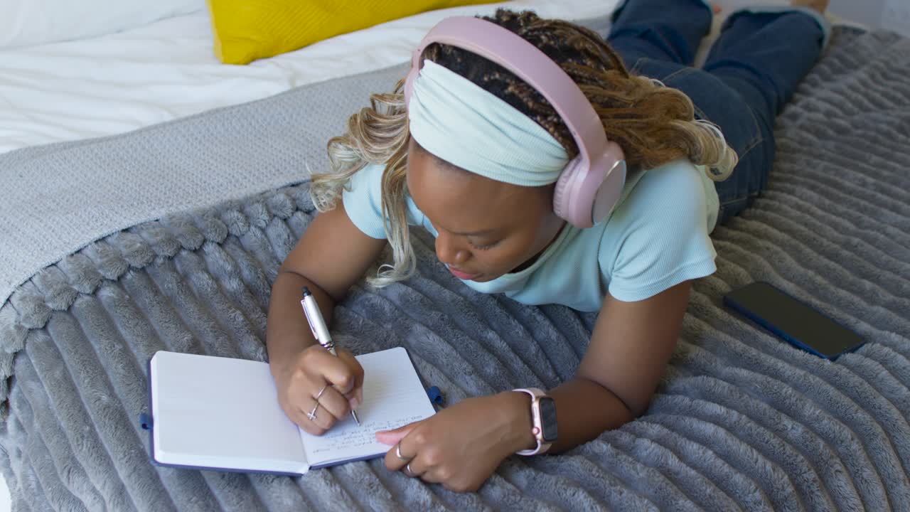 African American woman lying on bed with pink headphones using phone while taking notes in notebook