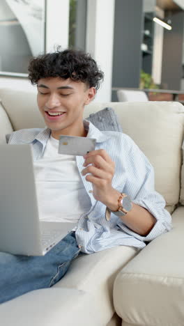vertical video: smiling young man shopping online with laptop and credit card at home
