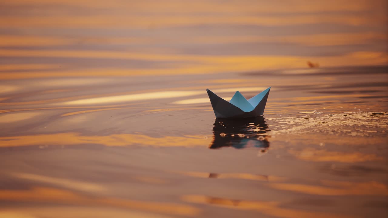 Origami boat on evening water background. Hand lets paper boat down into the water at sunset. Close-up.