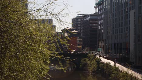 Sunlit-view-of-a-Manchester-canal-with-modern-buildings,-trees-and-clear-sky,-daytime