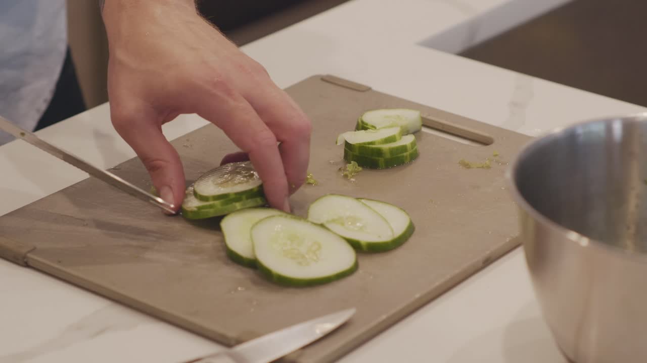 A closeup of cucumbers being sliced on a cutting board in a classy kitchen in preparation for a salad. Shot in Slow motion
