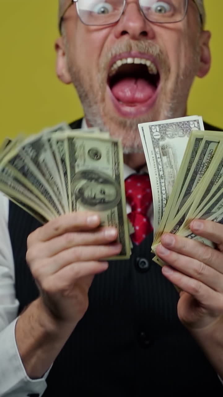 Excited mature man waving dollars. Happy funny elderly businessman showing banknotes on camera with opened mouth. Positive emotions. Vertical video