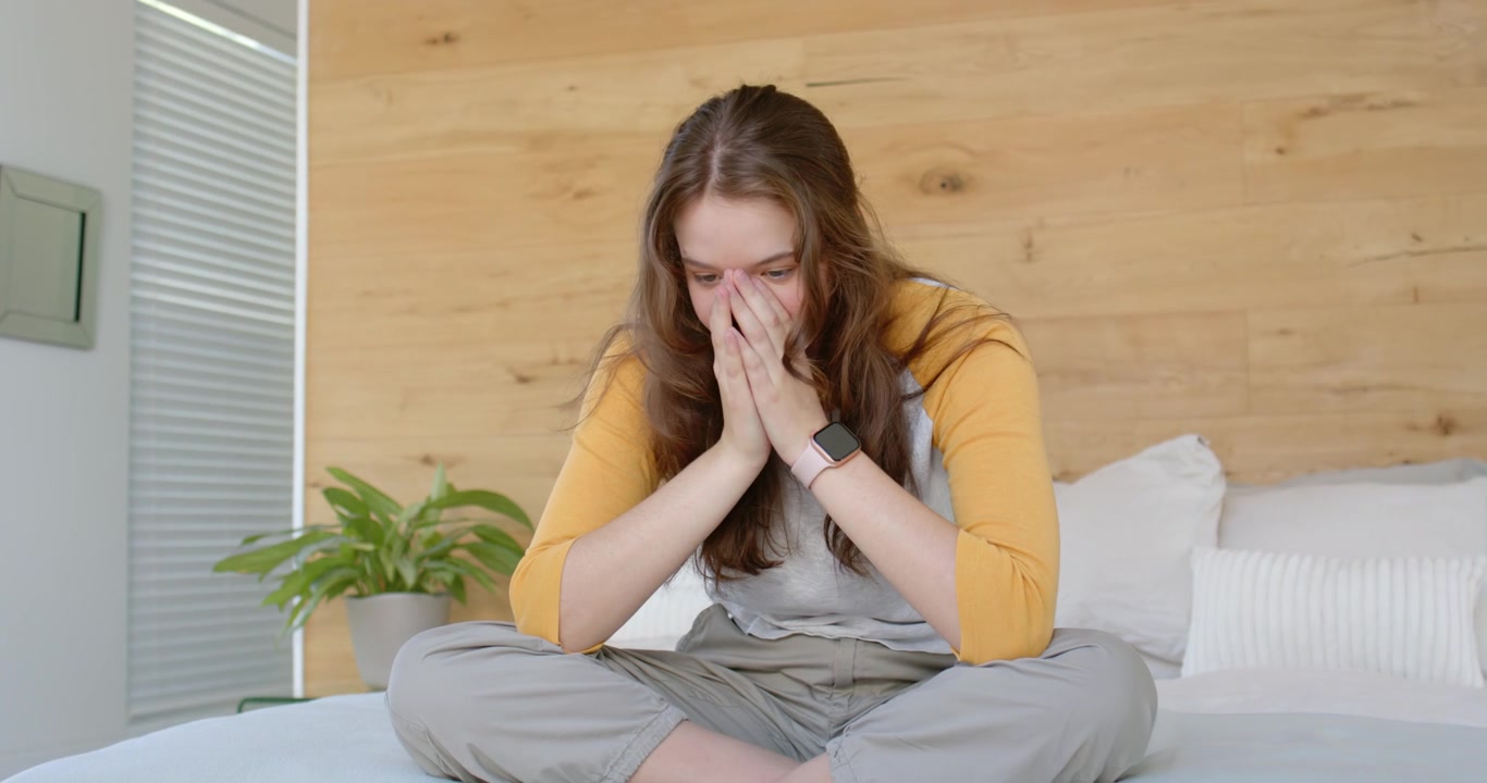 Donna seduta a gambe incrociate sul letto con le mani su bocca e naso, che tocca lo smartwatch