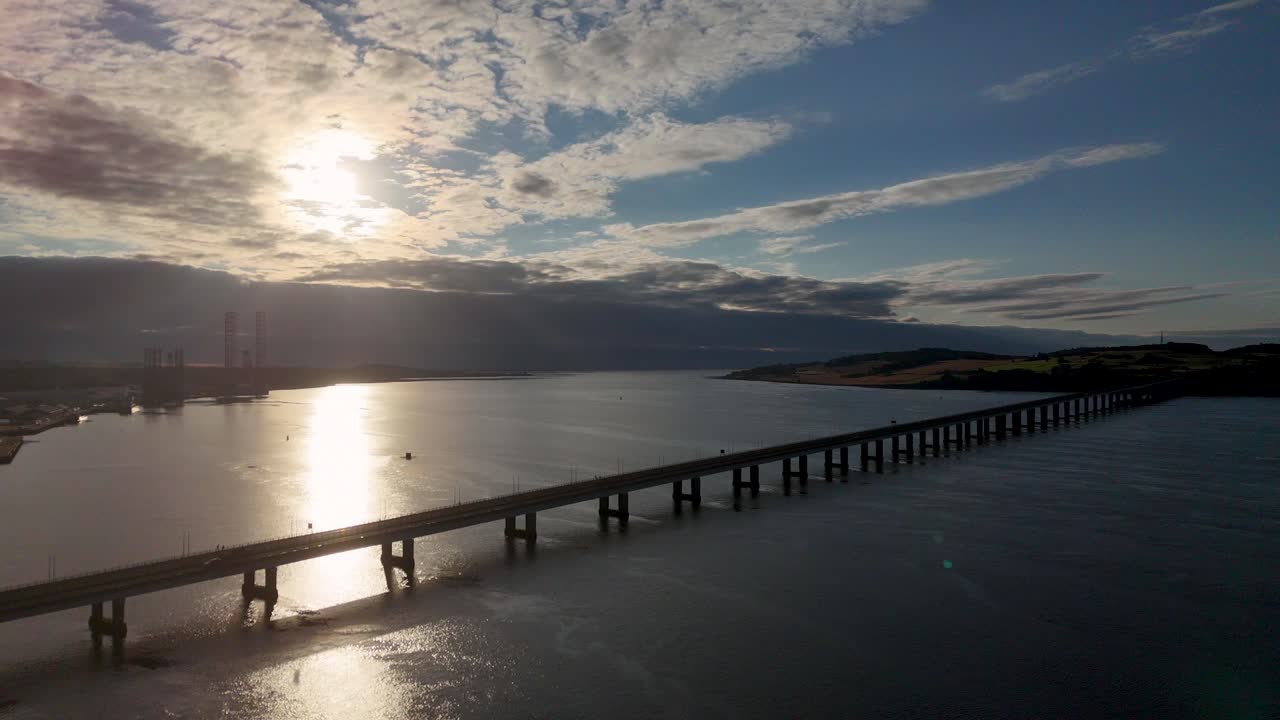 Aerial footage glides above a long bridge spanning calm water at sunrise in Inverness, Scotland, with dramatic clouds and golden reflections