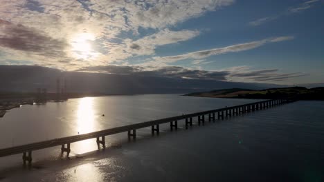 aerial footage glides above a long bridge spanning calm water at sunrise in inverness, scotland, with dramatic clouds and golden reflections