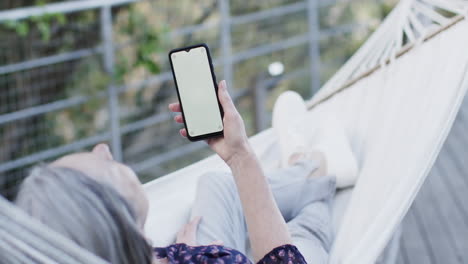 Kaukasische-Frau-Mittleren-Alters,-Die-Ihr-Smartphone-Benutzt-Und-In-Der-Hängematte-Auf-Der-Terrasse-In-Der-Natur-Entspannt,-Zeitlupe