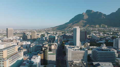 Forwards-fly-above-contemporary-office-buildings-in-city-centre.-Residential-neighbourhood-rising-up-on-slope-of-mountains-in-background.-Cape-Town,-South-Africa