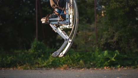 close-up of legs wearing dark clothing and metallic spring stilts, jumping on a paved path. the stilts showcase an intricate metallic design, and the background is a blurred green park