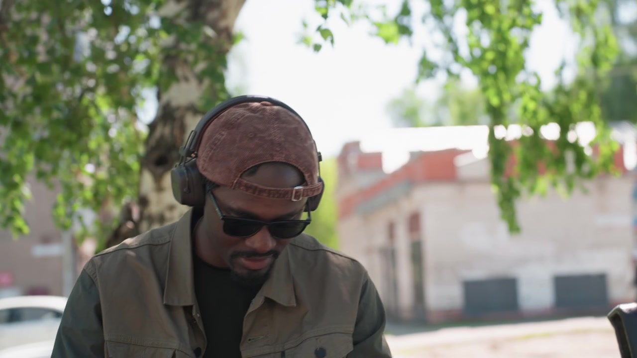Primo piano di un uomo con cuffie e giacca, seduto su una panchina e che tiene in mano il telefono in un parco con sfondo di alberi verdi. Occhiali da sole, cappellino, abbigliamento casual, all'aperto, luce diurna.