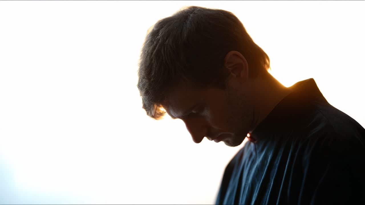 Un momento contemplativo catturato in silhouette, che ritrae un giovane uomo assorto nei suoi pensieri, con una luce soffusa che illumina i suoi lineamenti su uno sfondo sereno