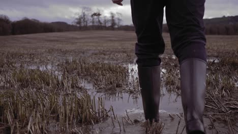 Hombre-En-Botas-De-Agua-Caminando-En-El-Campo-De-Tierras-De-Cultivo-Charcos-Fangosos,-Tiro-Bajo-En-Cámara-Lenta