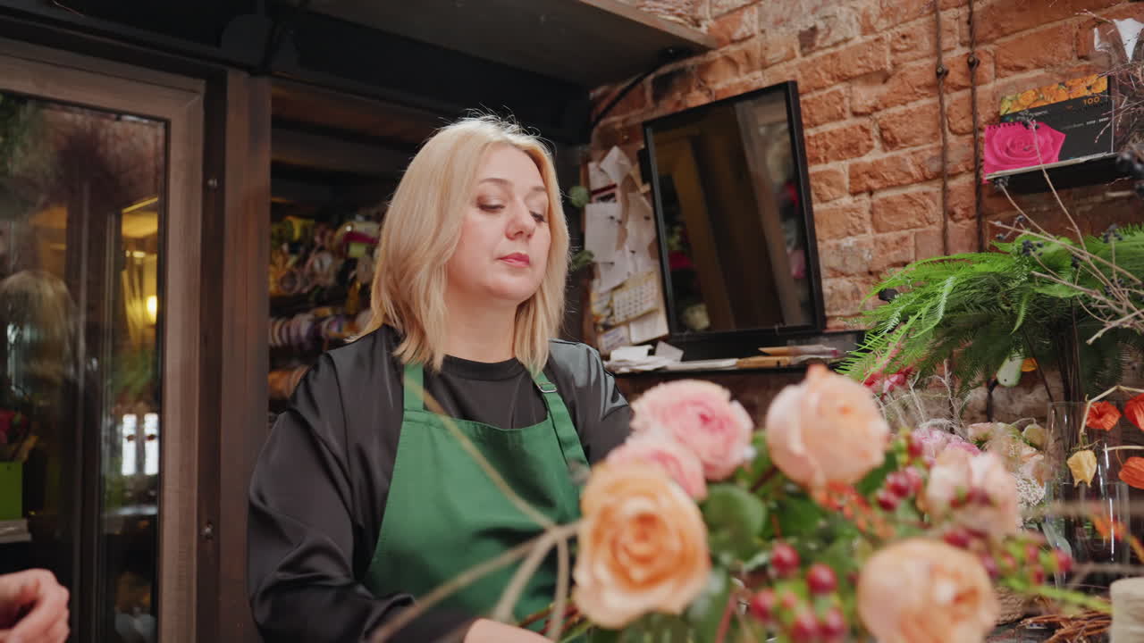 Fiorista bionda prepara un bouquet di rose color pesca in un laboratorio rustico in mattoni, selezionando i gambi, tagliando le foglie, avvolgendo il bouquet con carta artigianale e spago. Design floreale pratico, calda illuminazione ambientale, vasi.