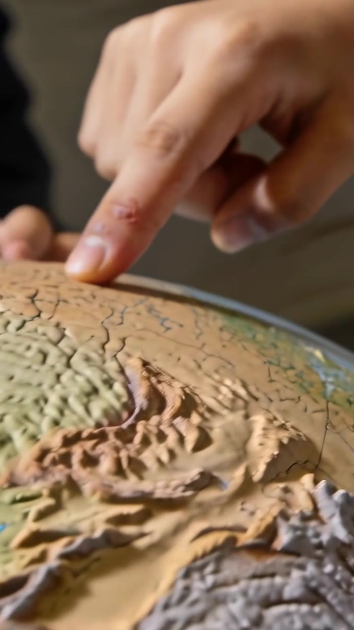 Video verticale: Mano con manica scura che preme il dito indice, esaminando un mappamondo in rilievo su un tavolo e tracciando una mappa.