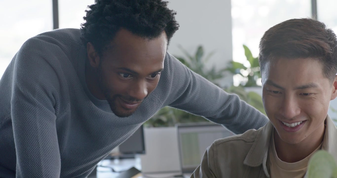 Diverse male team at desk, Asian coworker gesturing, African colleague rising, reviewing screen