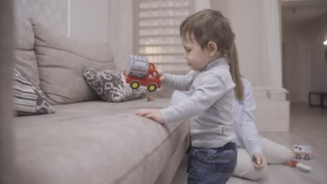 baby taking his first steps near the sofa to take a fire truck toy and give it to his mother. his mother helps him while he walking