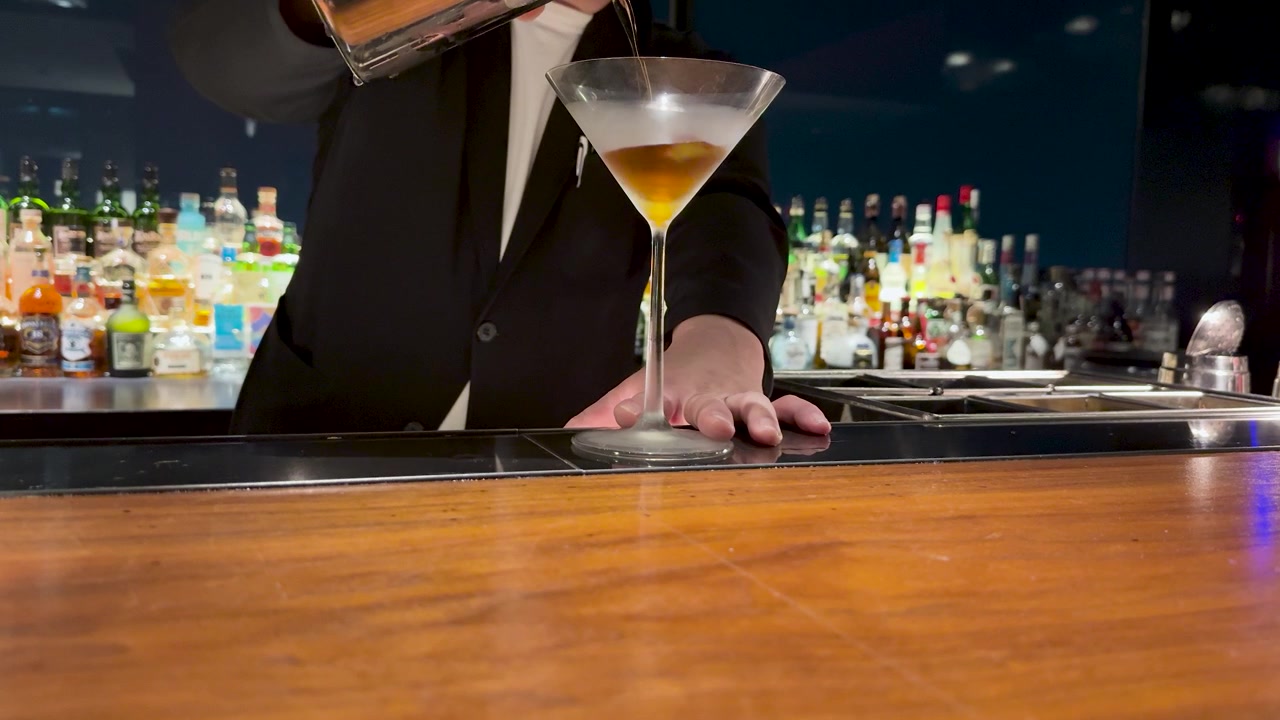 En professionel bartender hælder en mørk cocktail i et afkølet martiniglas på en bar.