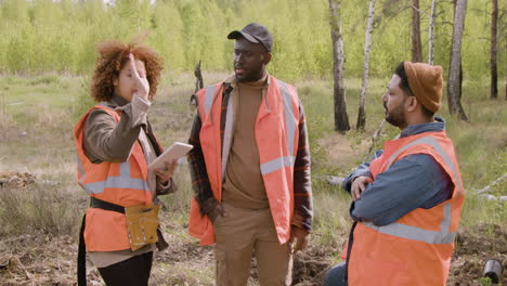 ativista afro-americana segurando um tablet e conversando com seus colegas de trabalho na floresta enquanto eles decidem onde plantar árvores